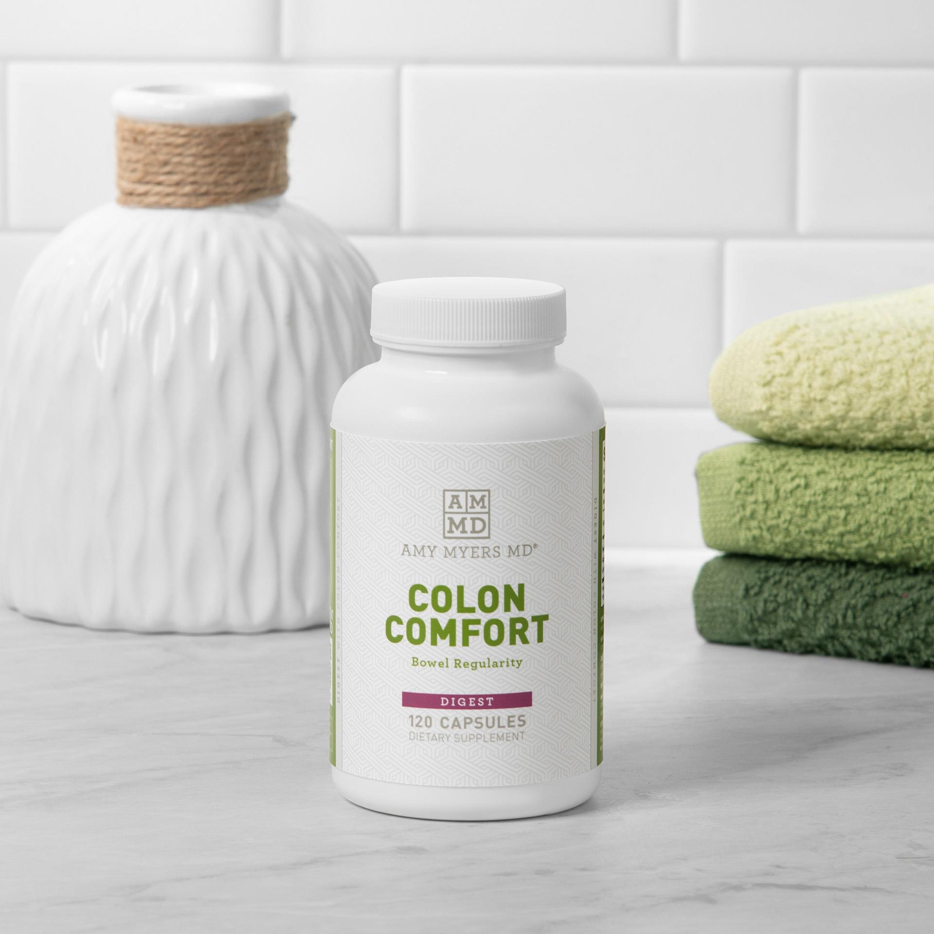 120 count capsule bottle of Colon Comfort sitting on a gray granite countertop with green colored towels, white vase, and white tile background.