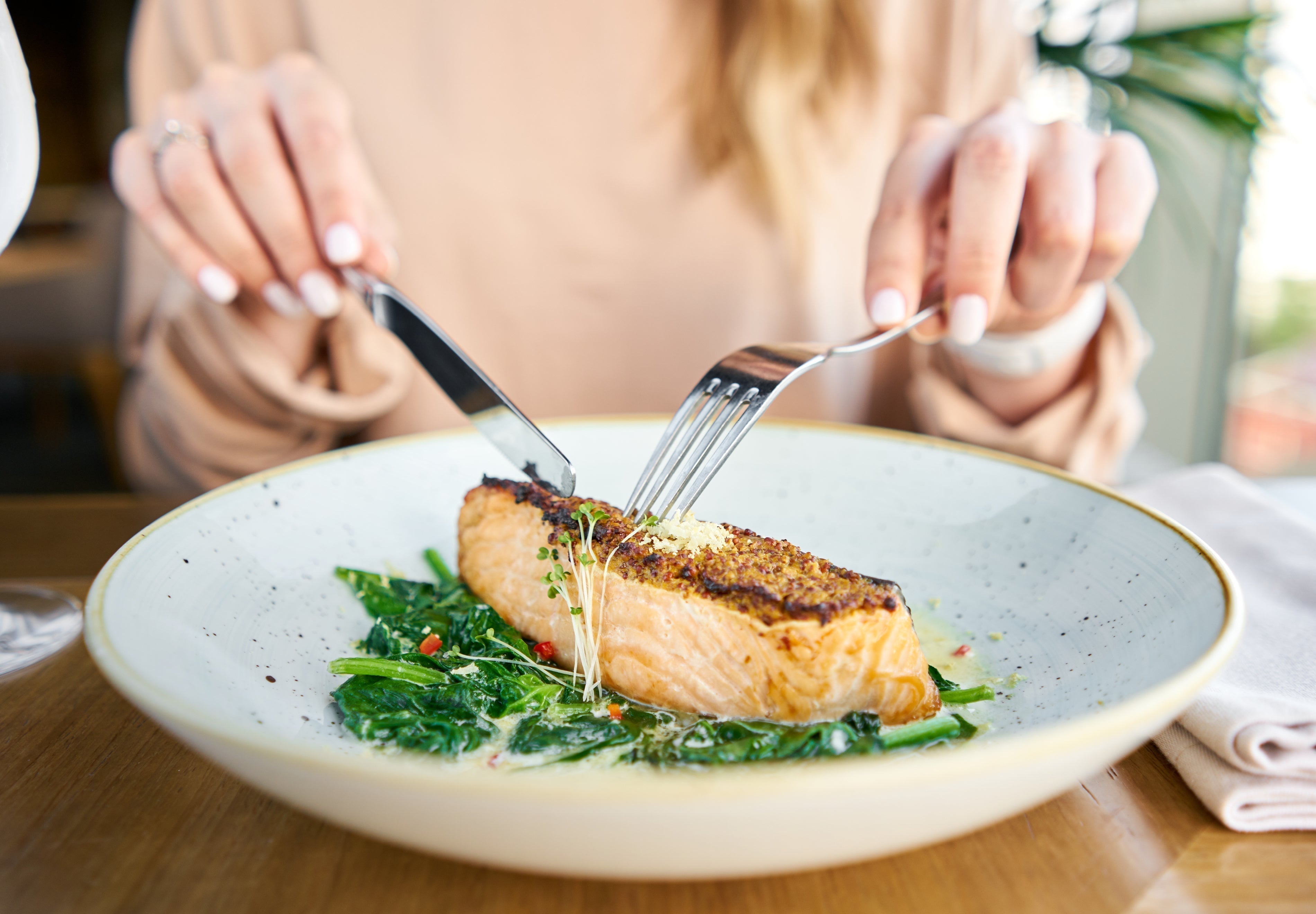 Person cutting into a seared salmon fillet served over sautéed greens at a restaurant table - Hypothyroidism Diet: Best and Worst Foods for Thyroid Health | AMMD™