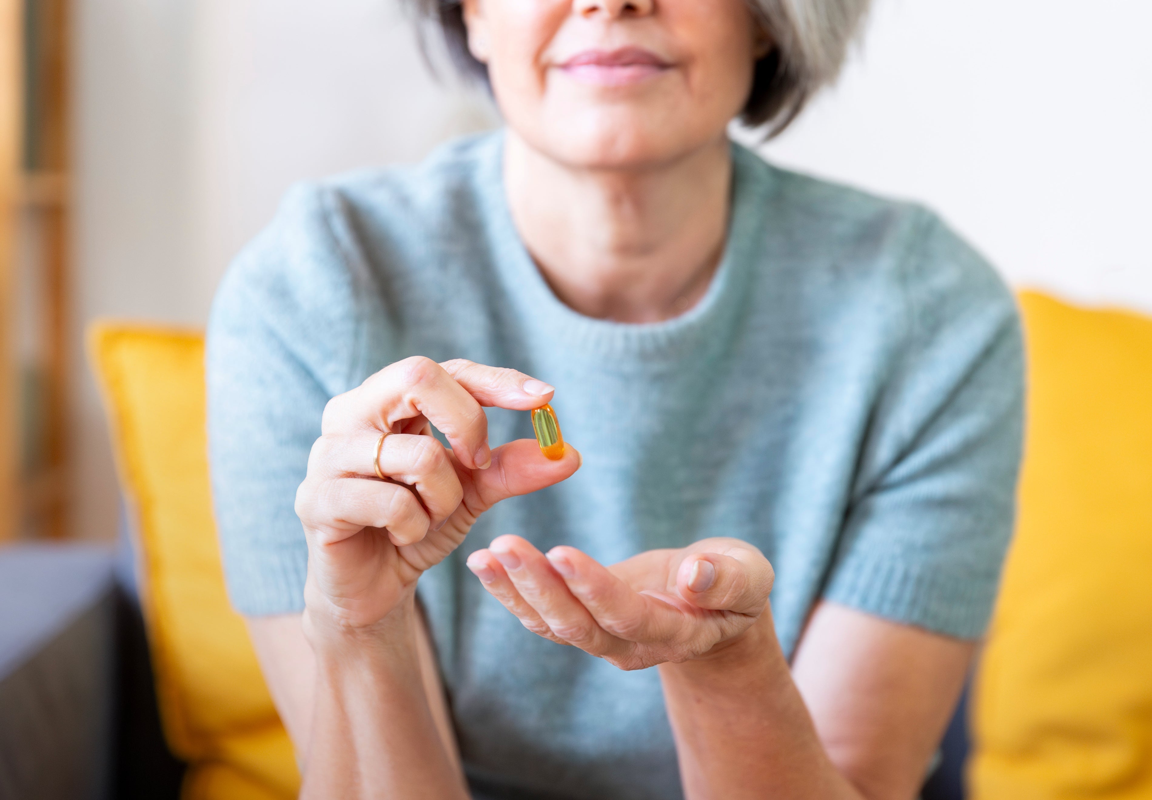 Woman holding a supplement capsule between her finWoman holding a supplement capsule between her fingers while sitting on a couch.gers while sitting on a couch.